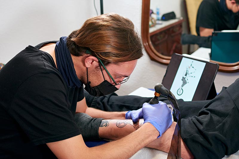 tattoo-artist-blue-gloves-looking-tablet-doing-tattoo-leg-male-studio artist giving someone a tattoo